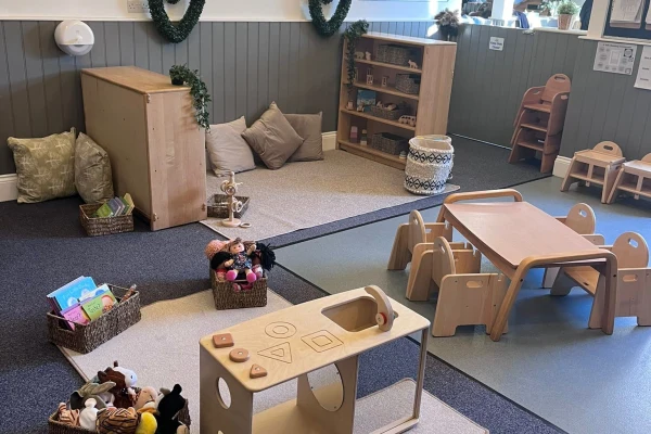 Low wooden play table and chairs sit beside a wooden activity cube; baskets of dolls and books are arranged on carpets near cushions and shelves in a calm preschool classroom.