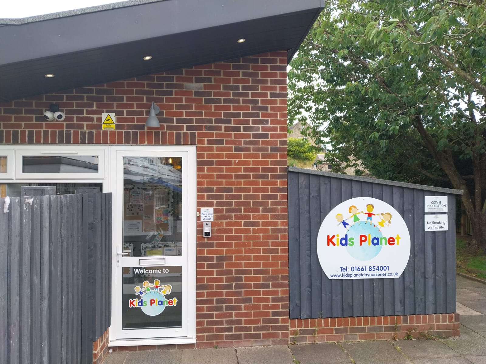 A nursery entrance: a white glass door and adjacent round "Kids Planet" sign on a grey fence; brick building frontage, security cameras, and a tree-lined sidewalk.