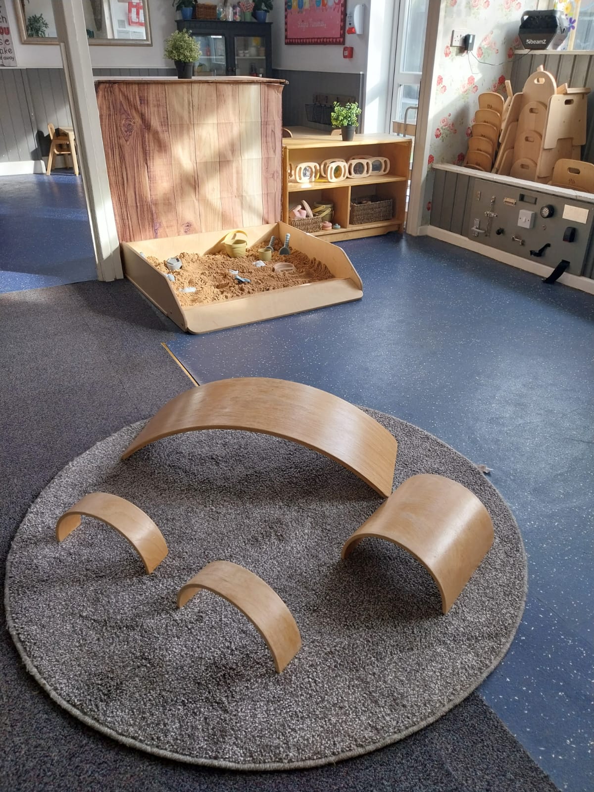 Wooden curved play planks arranged on a round gray rug, positioned for climbing and balancing inside a brightly lit children's playroom with a sandpit, low shelves, stacked chairs, and sunlight.
