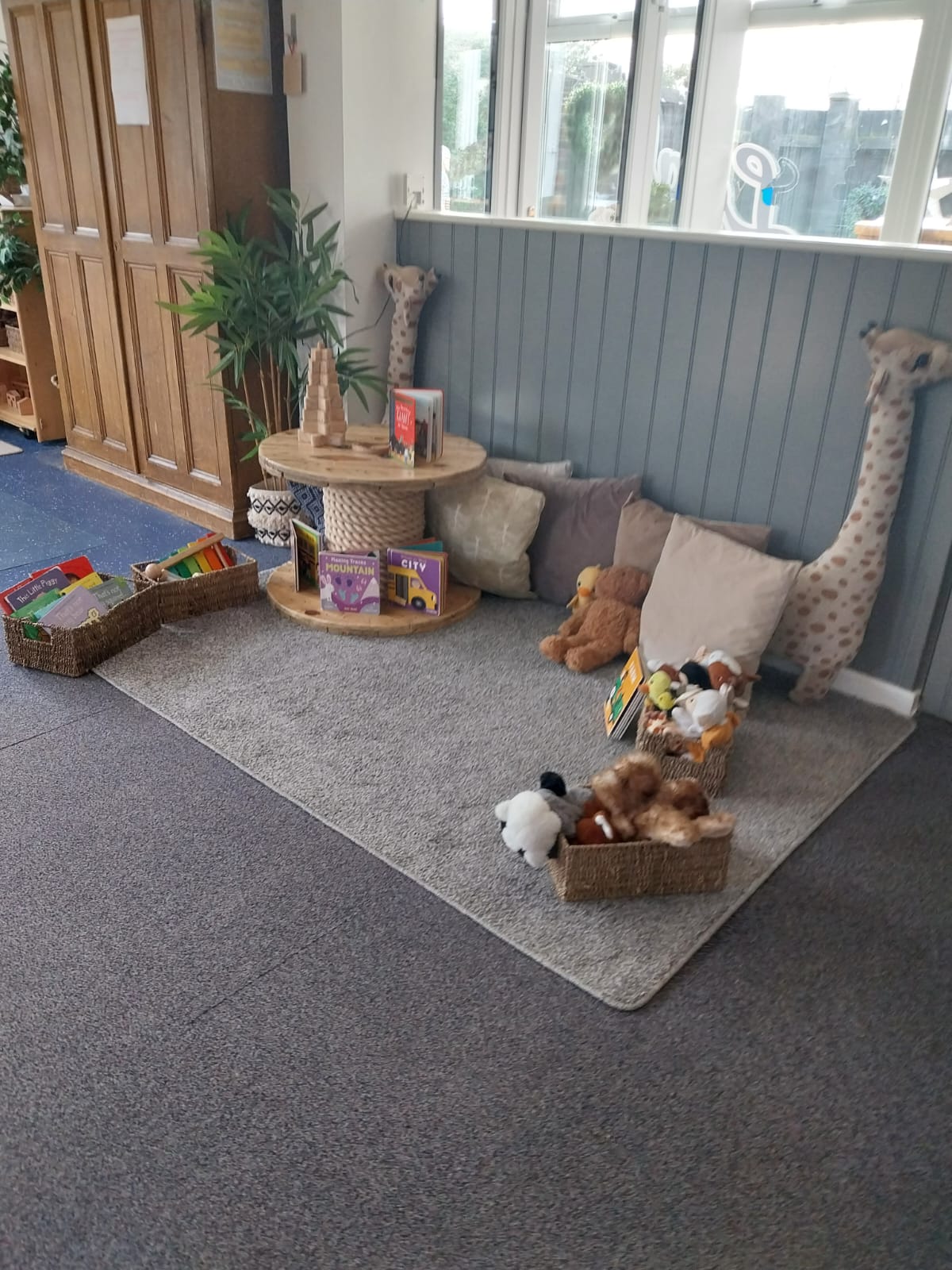 Children's reading nook: cushions and stuffed animals on a rug, baskets of books and toys beside a small wooden table under windows and a grey beadboard wall.