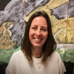 Woman with shoulder-length brown hair smiles at the camera, wearing a light sweater and necklaces, standing before a mural of large yellow birds and gray brushstrokes over a green band.