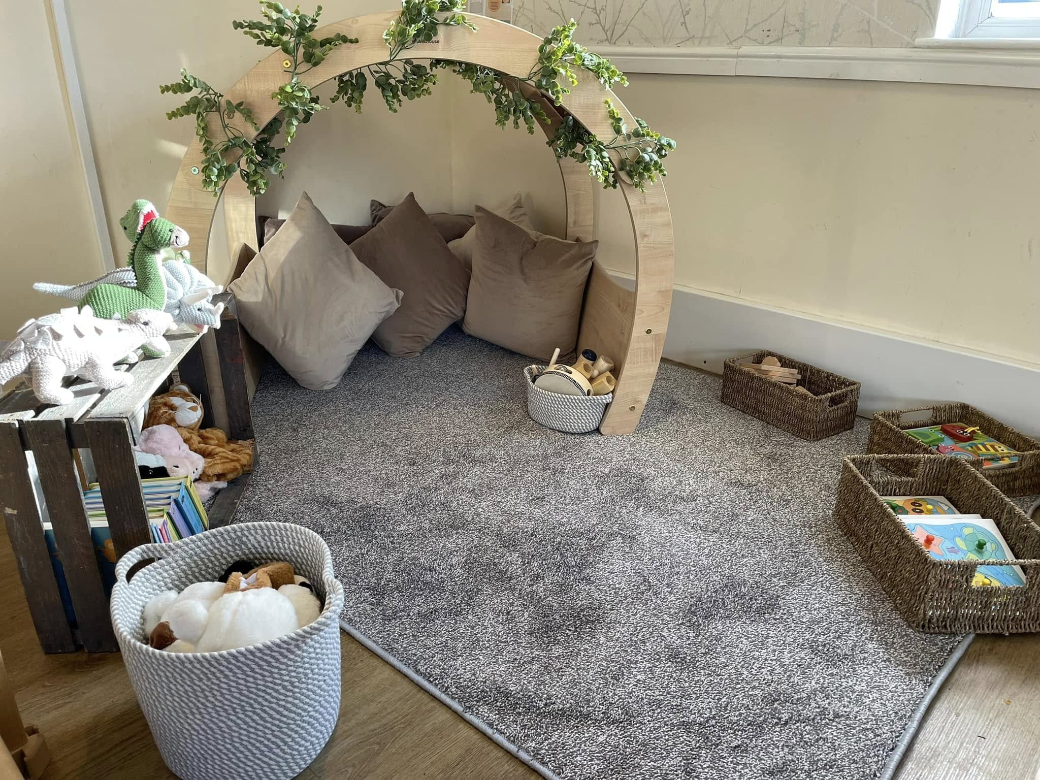 Wooden arch framing cozy reading nook; neutral cushions tucked beneath. Soft gray rug anchors corner play area with woven baskets, stuffed animals, books and sunlight from a window.