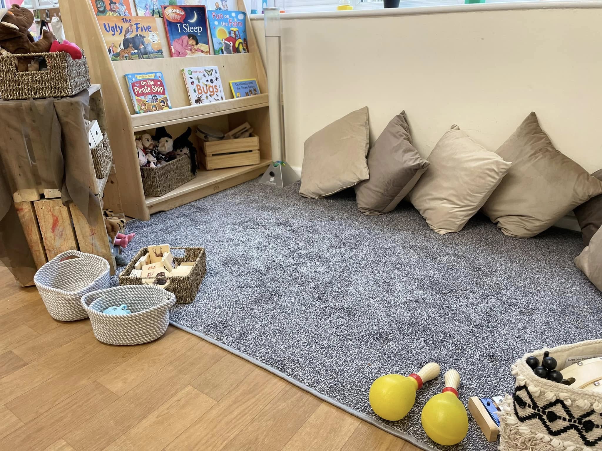 Gray carpeted reading area with beige cushions against a low wall; woven baskets and wooden toys, including two yellow maracas, sit on the floor beside a small bookshelf in a classroom corner.