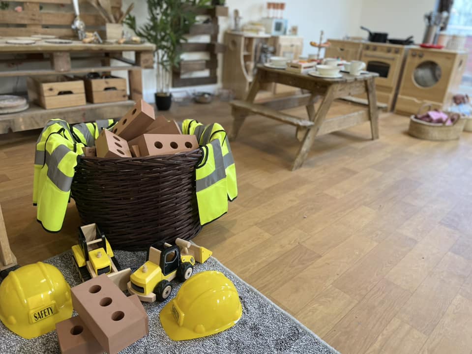 Wicker basket of toy bricks draped with hi‑vis vests; sits on a wooden floor while toy construction vehicles and two yellow hard hats (one labeled "SAFETY") rest nearby in a children's playroom.