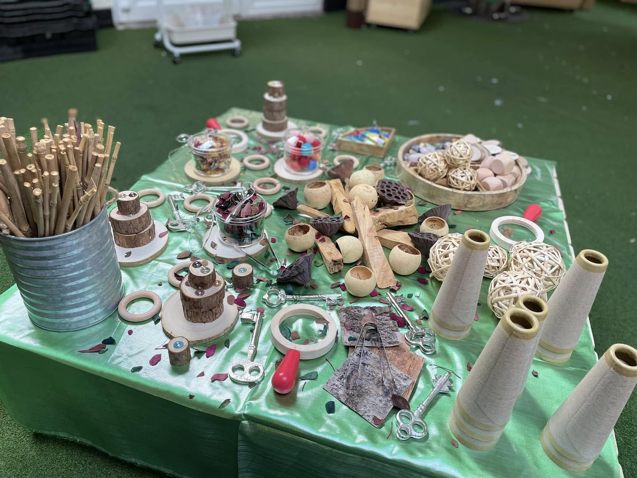 A craft-materials table displaying wooden discs, sticks, woven balls, magnifying rings and keys; pieces arranged scattered for activity on a green turf floor in an indoor play area.