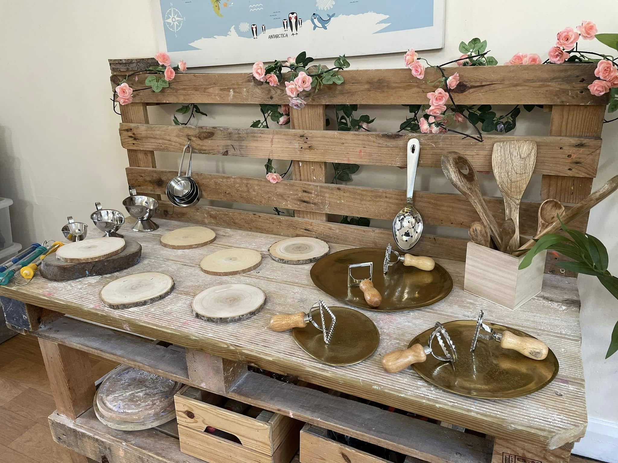 Wood slices and brass plates with wooden-handled pastry crimpers are arranged on a rustic pallet workbench, surrounded by wooden utensils, hanging measuring cups and pink artificial flowers; map text: "ANTARCTICA".