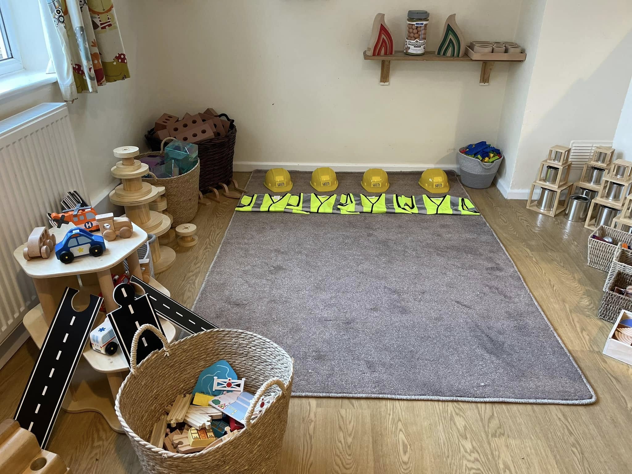 Four yellow helmets labeled "SAFETY" sit in a row with high-visibility vests on a carpeted playroom floor; wooden toys, baskets and shelves surround the space, toy helicopter marked "H".