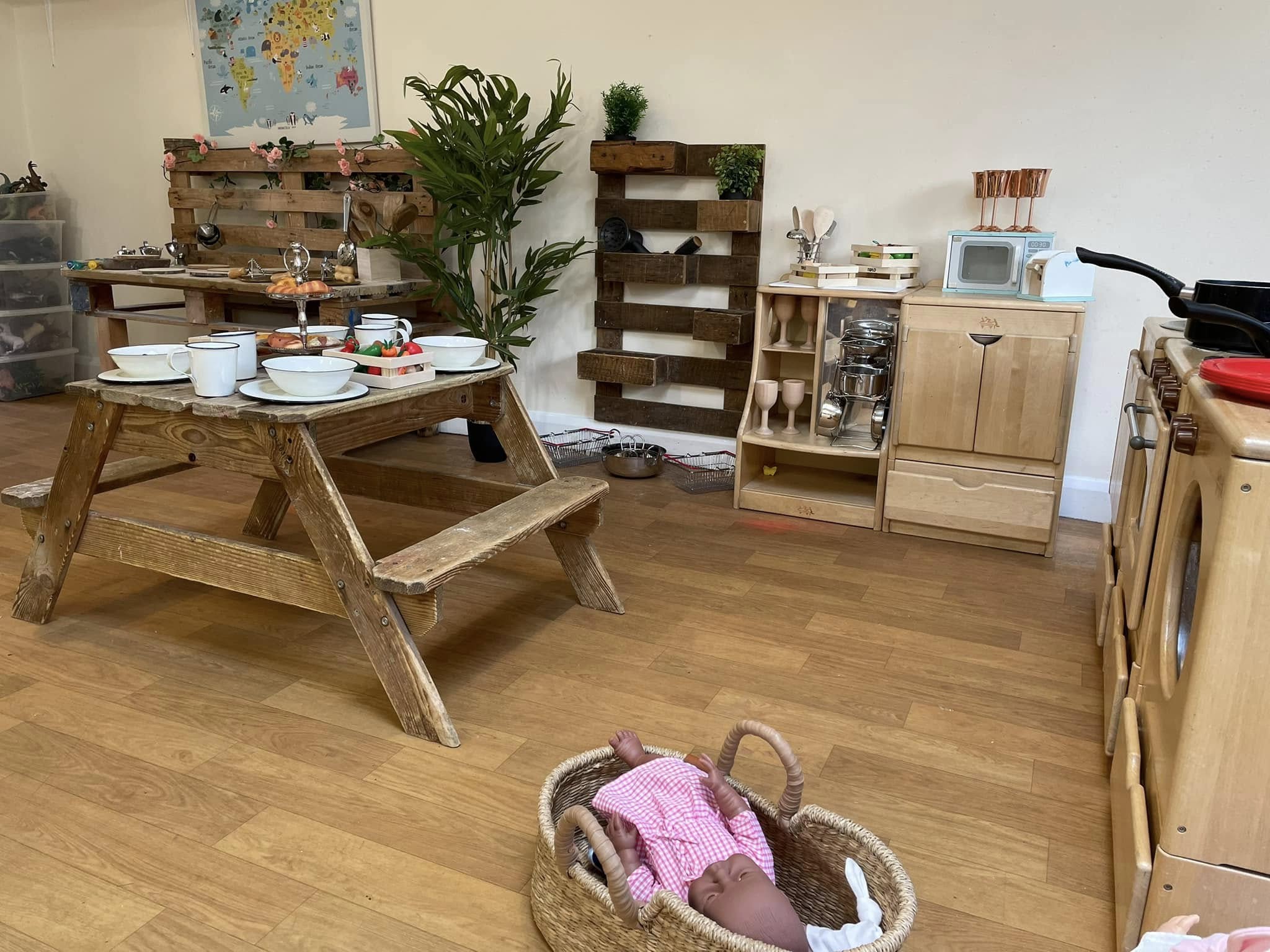 Wooden picnic table holds bowls, mugs and toy food arranged for a meal; inside a children's playroom with play-kitchen, pallet shelves, plant, and a doll in a woven basket.