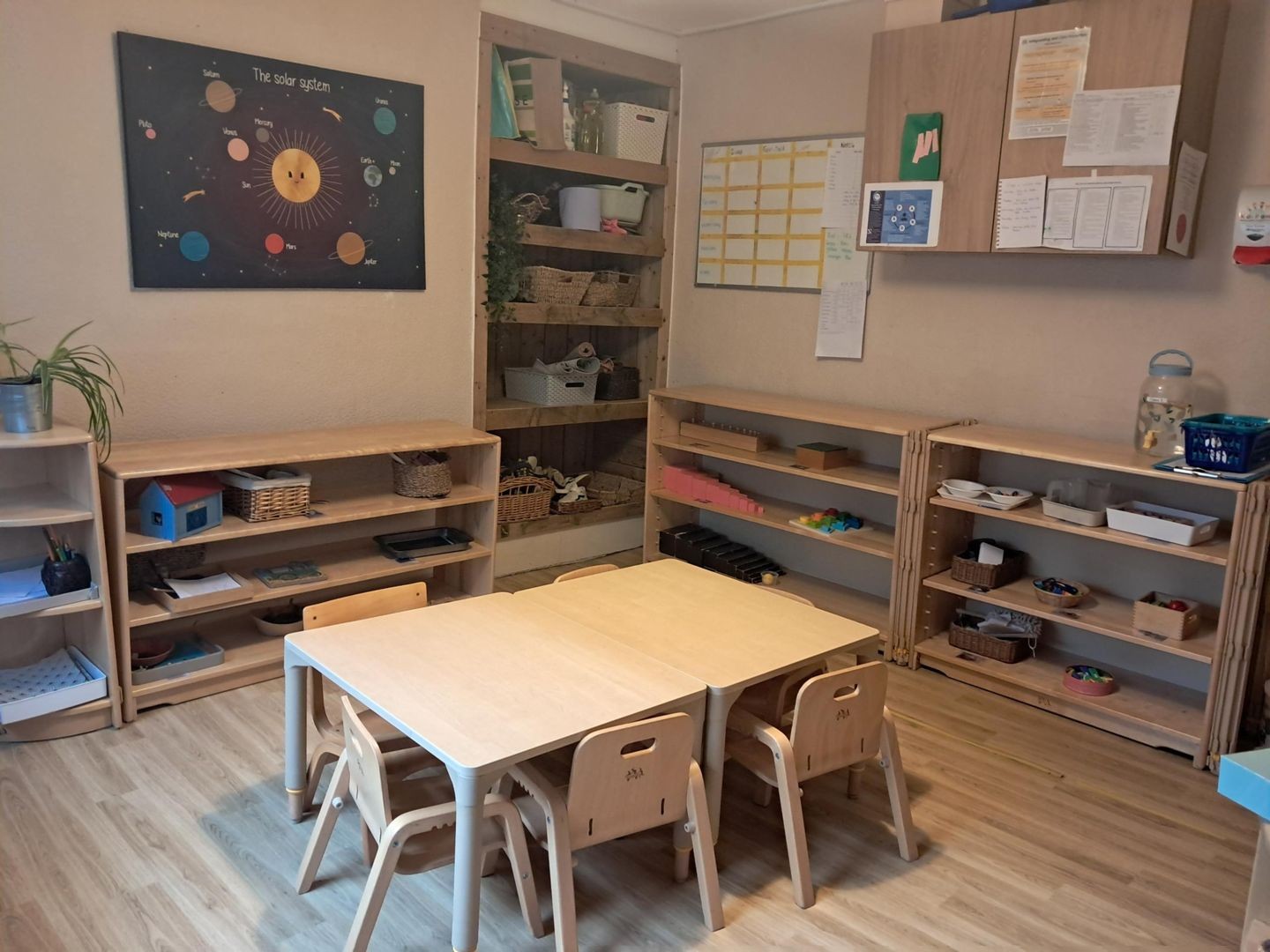 Small wooden children's tables and four chairs arranged for activity, surrounded by low shelves with baskets, toys and a smiling solar-system poster on the classroom wall; laminated chart and cabinets above.