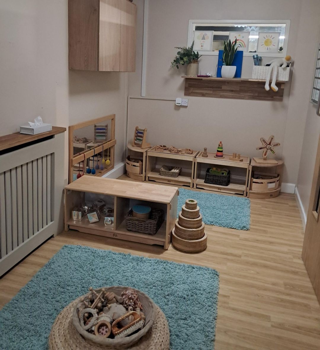 Wooden toys and shelves arranged neatly across a small children's playroom — toys stacked and placed in baskets — with two blue rugs, wood flooring, a wall shelf holding plants and art.