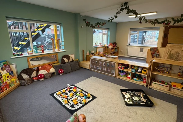 Children's playroom: low shelves and baskets hold books, toy vehicles, blocks and cushions; two play mats with a road set and building blocks lie on carpet beneath windows and garlands.