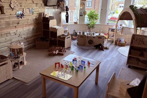 A low wooden table displays colorful translucent blocks arranged on small mirrors, set in a sunlit children's playroom filled with wooden shelves, a toy kitchen, rugs, and large front windows.