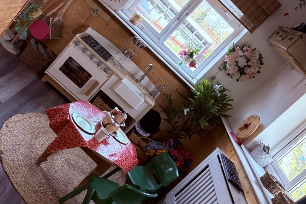 Children's wooden play kitchen and small table set with toy dishes arranged, surrounded by green plastic chairs, a rug, potted plants, window light, and decorative wreath in a cozy playroom.