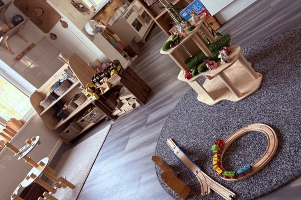 Wooden toy train on a circular track sits on a gray rug; surrounded by tiered wooden play tables, low shelves with baskets, and natural light in a tidy Montessori-style classroom.