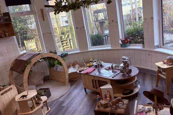 Wooden play furniture and toys occupy a children's play area, arranged around a low play kitchen and reading nook beneath hanging plants in a bright bay-windowed room overlooking a garden.