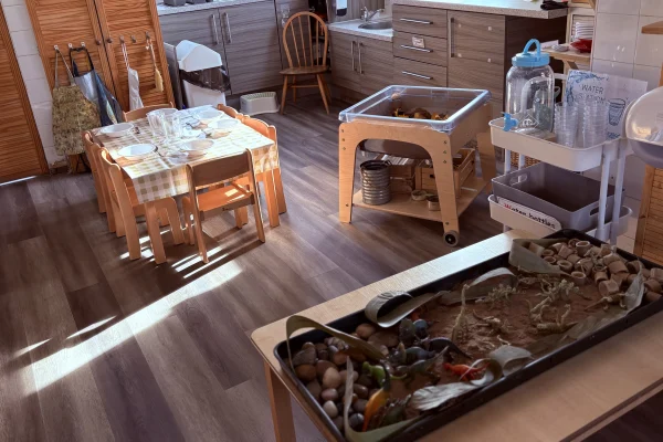 Children's table set with plates and cups arranged for a pretend meal, bathed in sunlight, beside sensory play trays and a small wooden play kitchen with cabinets. Text: WATER.