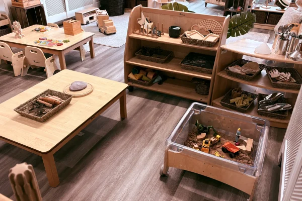 A low wooden sensory bin holds toy trucks in sand, surrounded by low tables and shelves displaying baskets of wooden toys and pretend food on laminate flooring in a classroom.