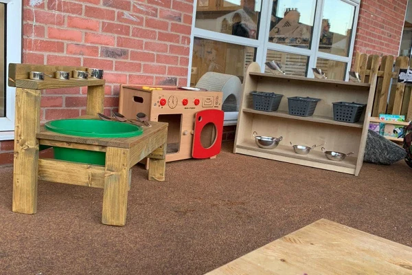 Wooden toy kitchen set (green basin sink, red toy oven, shelving with baskets and metal bowls) sits arranged on a brown outdoor play mat against a brick wall beneath windows.