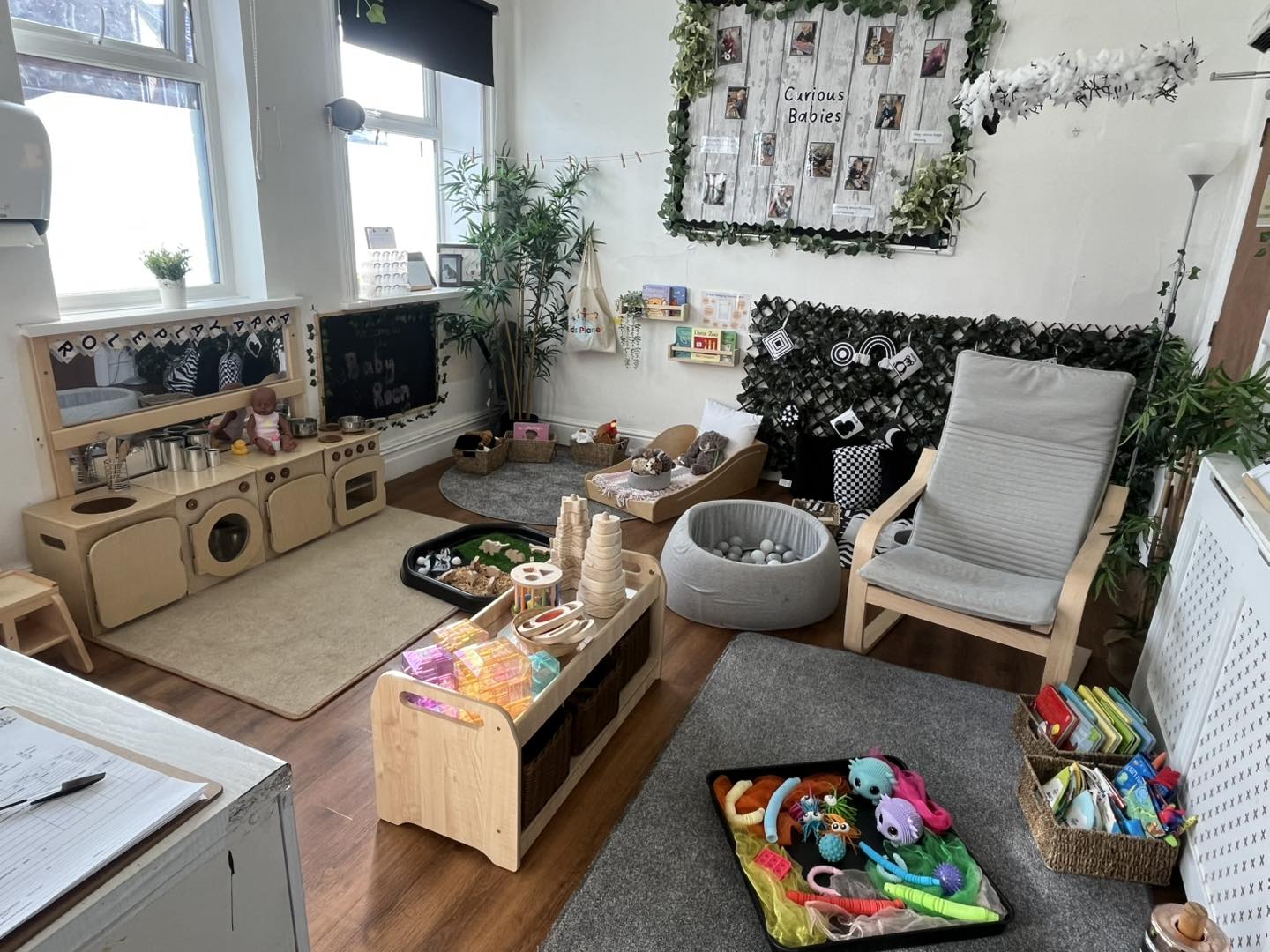 Children's play corner filled with wooden toys, soft seating, and activity trays, arranged beneath windows and a photo board, surrounded by plants, rugs, and cozy seating.