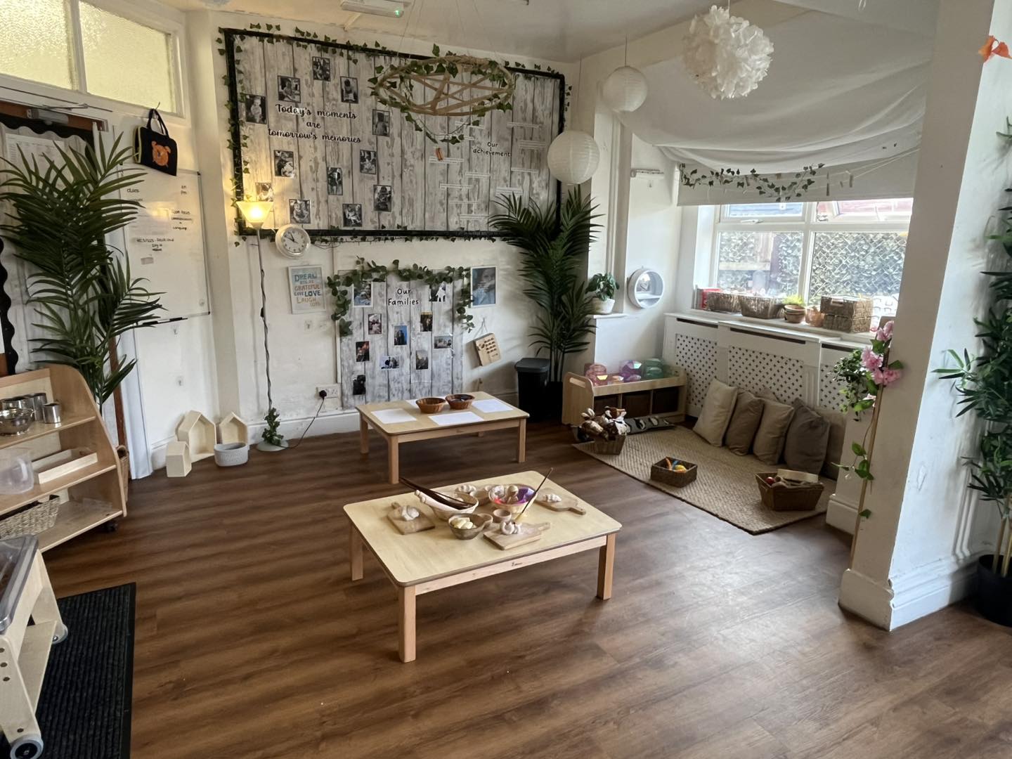 Low wooden table displays wooden bowls and pretend food in a cozy children’s play area with cushions, plants, hanging lanterns, and photo-filled bulletin boards by a sunlit window.