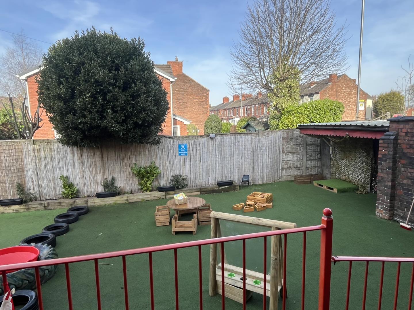 Small fenced children's play area holding a round wooden table, crates and scattered blocks and tires on green turf, backed by a bamboo fence and suburban brick houses.