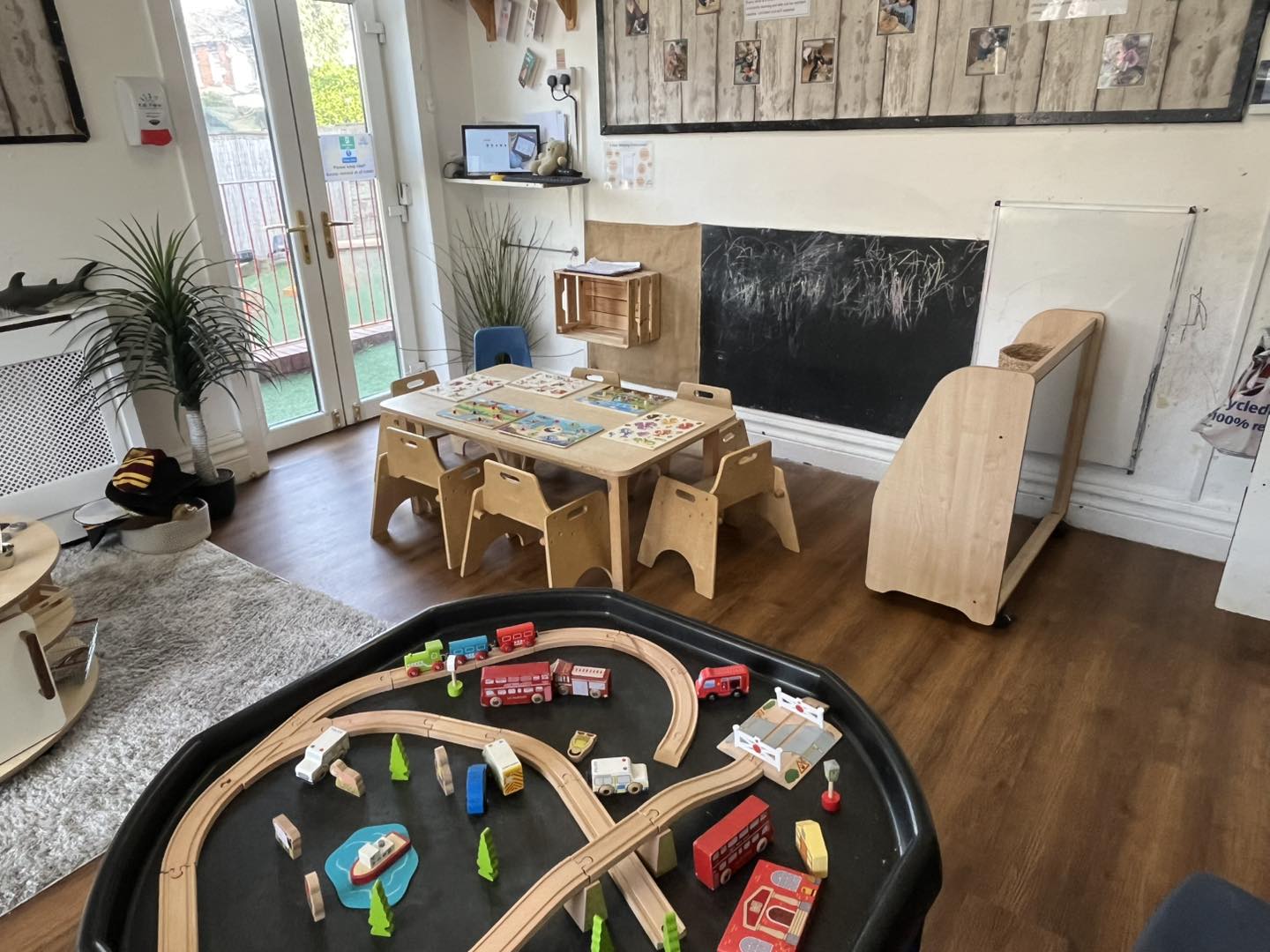 Wooden train set arranged on a black play tray, surrounded by toy vehicles and trees, in a bright preschool playroom with low tables, child chairs, chalkboard and double doors.