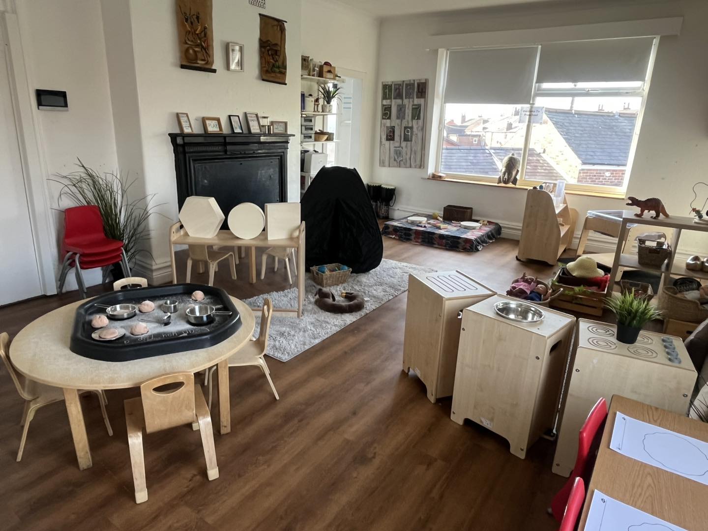 A low round wooden table holds a black sensory tray with dough, bowls and utensils; small chairs circle it in a bright classroom with play stations, rug, toys and a window.