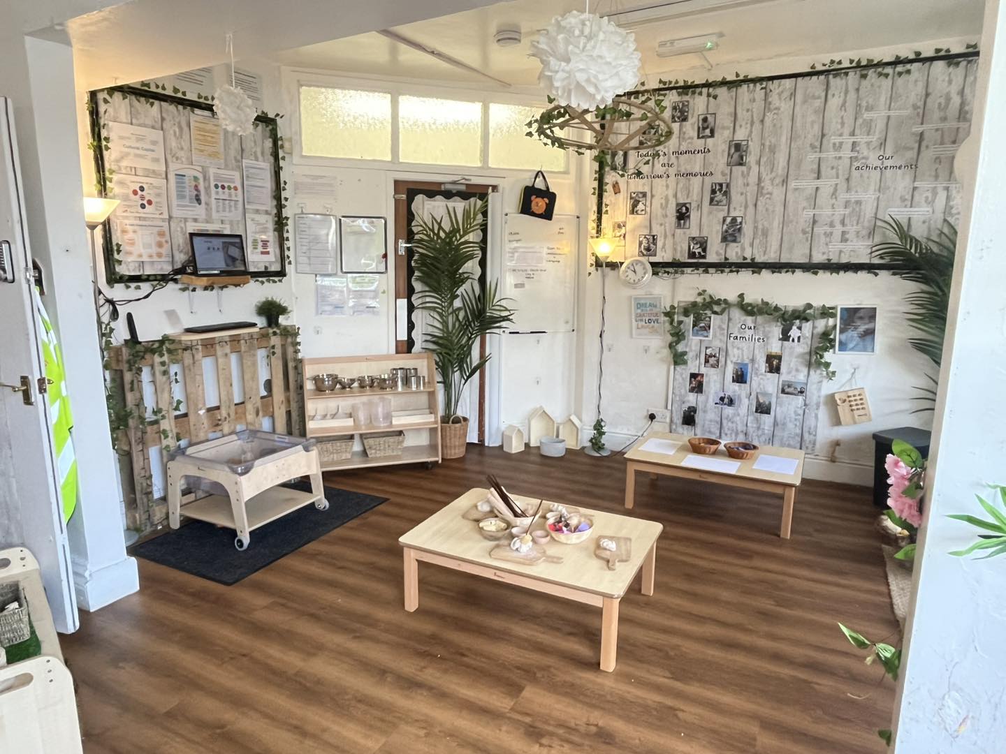 A low wooden table holds bowls of play materials; a second table and shelving sit on a wood floor in a bright, plant-filled early-years classroom with photo and info boards.