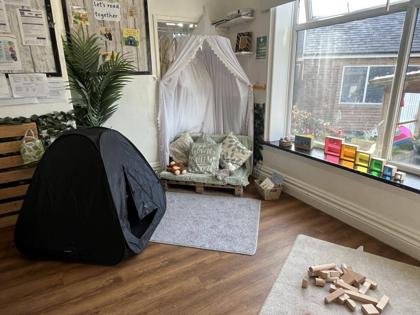 Black pop-up tent sits on a wooden floor; nearby a white canopy nook holds cushions on a pallet seat by a large sunlit window; wooden blocks scattered on a rug.