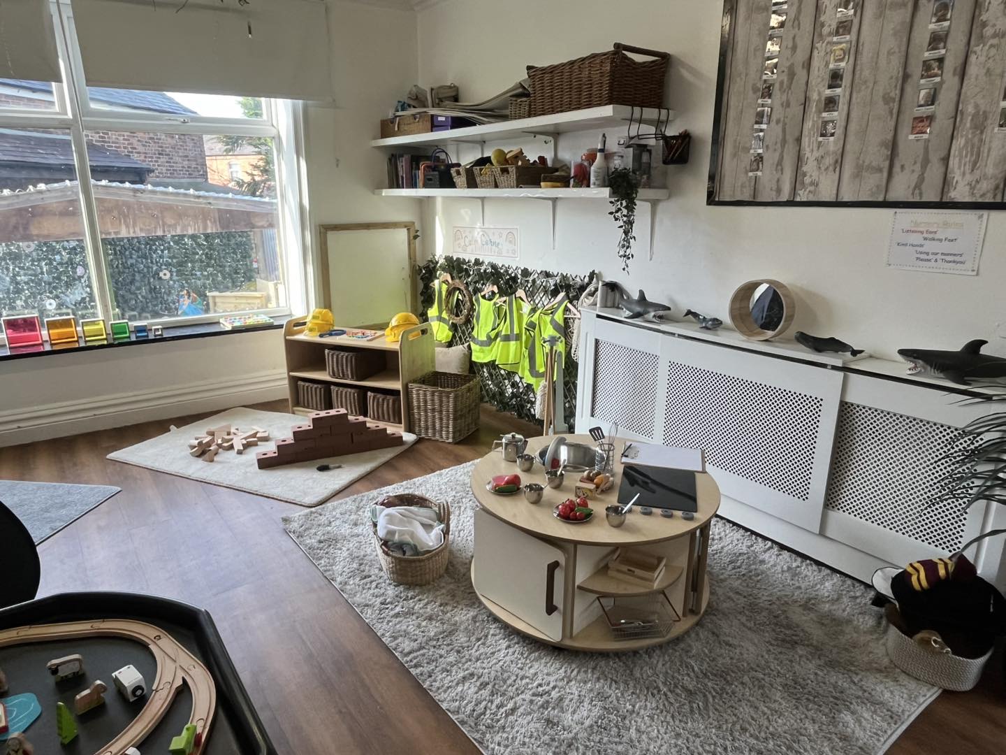 Low round play table holds toy kitchenware and pretend food, set on a rug in a sunlit preschool playroom with shelves, dress-up safety vests, building blocks, window and radiator display.