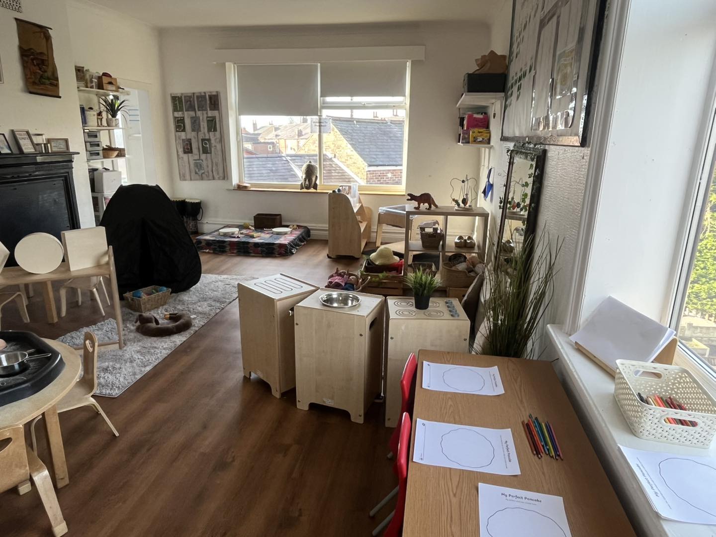 Child-sized table holds drawing sheets and colored pencils; red chairs beside it; play kitchen cubes, rugs and toy shelves fill a sunny room with a cat on the windowsill.