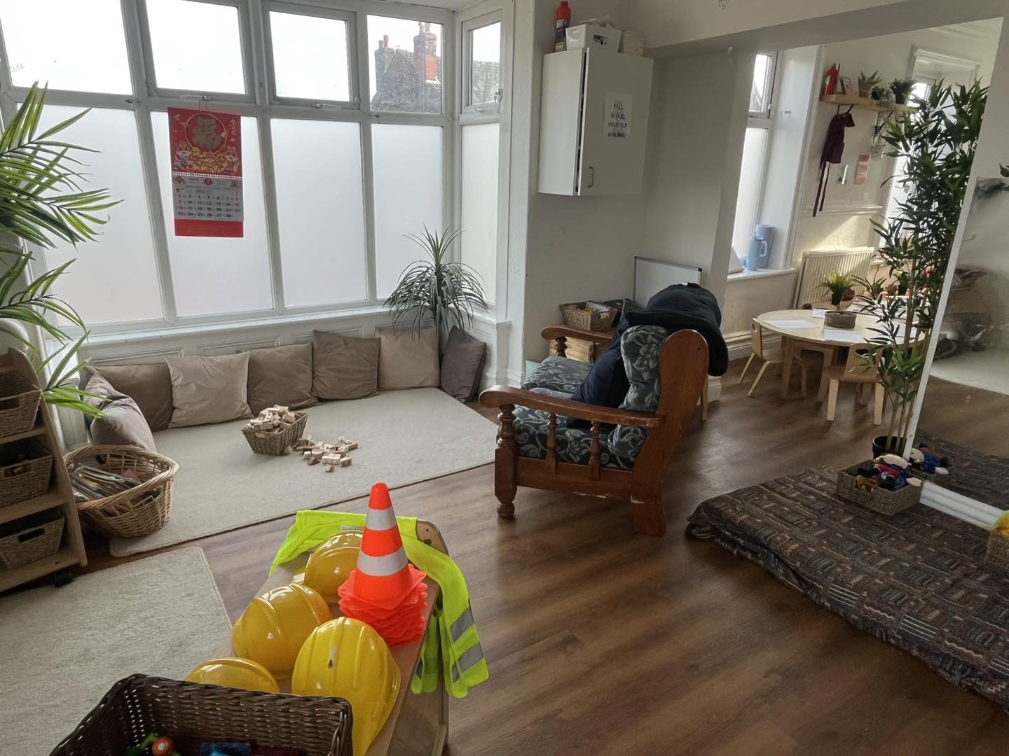 Yellow construction helmets stacked beside a neon safety vest and orange cone, in a sunlit children's playroom with low floor cushions, wooden toys, small table, and plants by frosted windows.