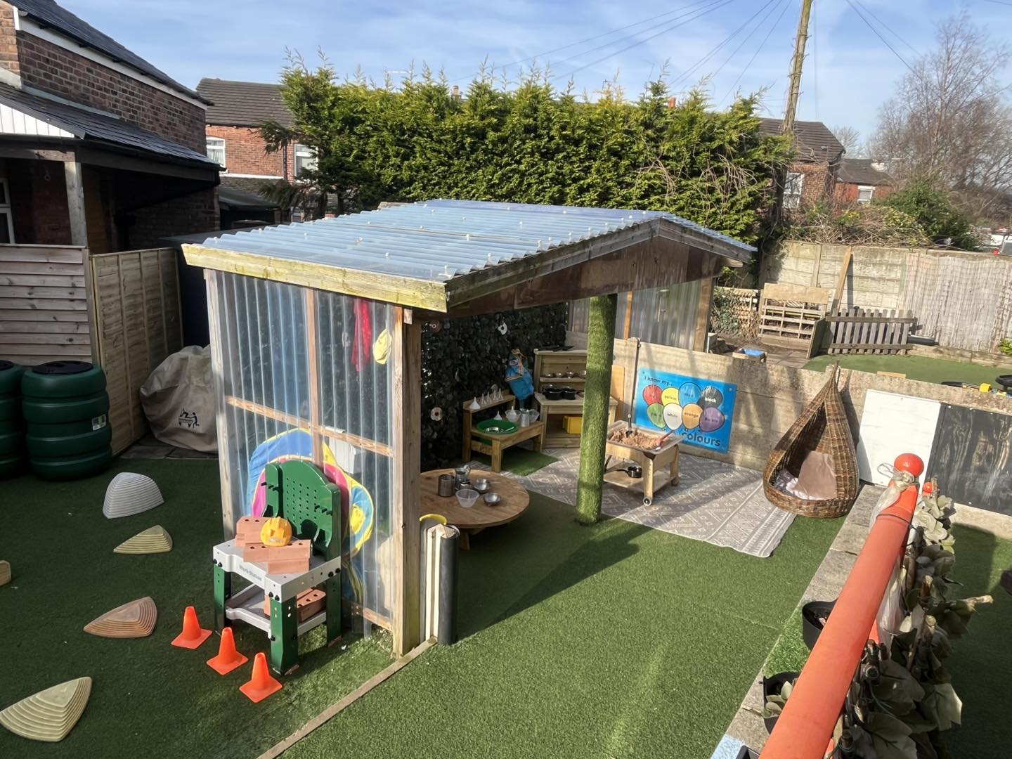 Wooden play shelter houses a toy kitchen and seating, providing shaded play space in an artificial-turf backyard with fences, a hanging wicker chair and nearby houses. Text: "Colours"