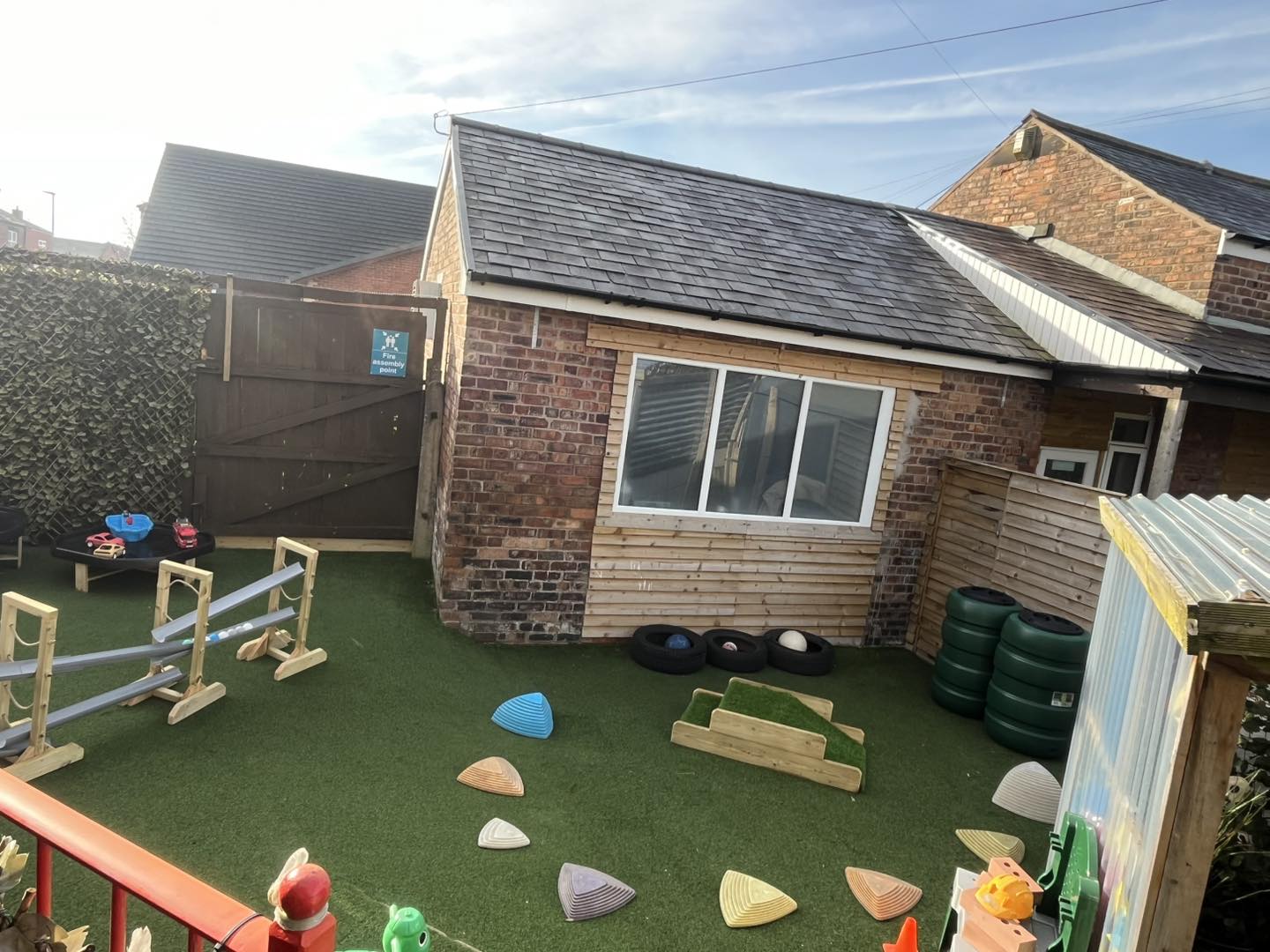 Backyard play area: colorful plastic stepping stones, wooden ramps and toy cars scattered on artificial turf, beside a brick outbuilding and wooden gate.