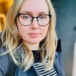 Person with black-rimmed glasses looks directly at the camera; wearing a black-and-white striped shirt, gray jacket and backpack strap, standing indoors against a softly blurred background.