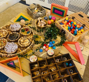 A wooden table displays sensory materials: wicker baskets and trays filled with colorful wooden balls, geometric frames, beads and blocks, arranged for children's play in a bright indoor classroom.