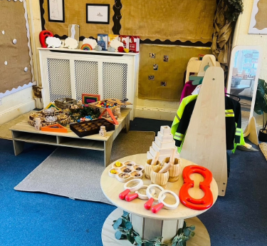 Round toy table displays nested bowls, blocks, utensils and red weights; low shelves hold sensory trays and loose parts; a coat rack with hi‑vis jackets marks a classroom corner.