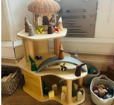 Wooden multi-level toy house with a wicker-roof top; peg gnome figurines stand and cross a small arch bridge while baskets of pastel eggs sit beside it on a hardwood floor. No text found.