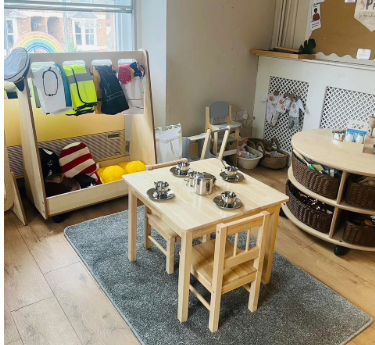 Small wooden children's table and chairs hold a metal toy tea set, arranged for pretend tea on a gray rug amid a playroom with dress-up rack, helmets, baskets, window rainbow.