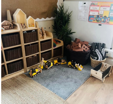 Wooden toy construction vehicles are lined up on a rug, beside cubby shelves, baskets, toy houses and a small wagon in a classroom corner. Text visible: "Our calendar", "Thursday", "Winter", "Monday".