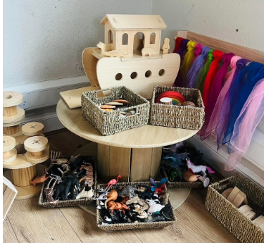 Wooden ark sits on a round spool shelf, surrounded by baskets of toy animals and accessories; colorful scarves hang on a low wall rack in a wooden-floored corner.