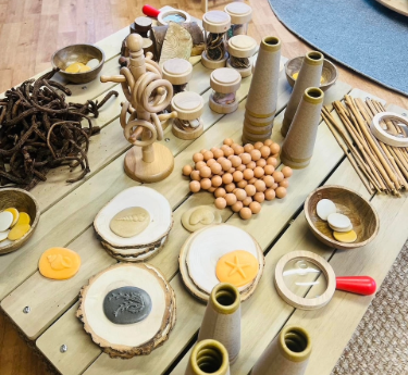 Wooden play materials — tapered cones, round beads, rings and disc slices — are arranged on a light slatted table, with bowls of discs and bundles of sticks in an indoor area.