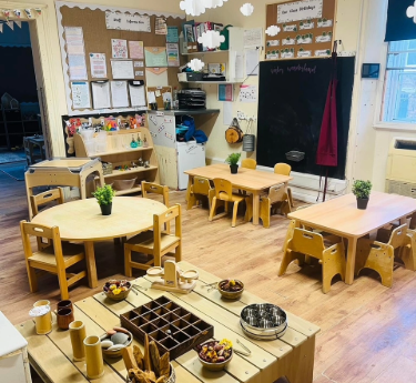 Small wooden tables and child-sized chairs arranged neatly, holding potted plants and craft materials; activity stations and shelves with toys fill a bright preschool classroom with a blackboard and displays.