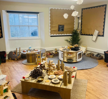 Wooden play tables display wooden toys and pretend food, arranged on rugs in a sunlit preschool corner with blank bulletin boards, a window, potted plant, and storage baskets. A small chalkboard sign on the wall has text too small to read.