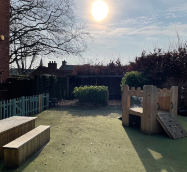 Wooden play structure sits atop artificial-turf yard, casting long shadow as low sun shines; tiered benches and small climbing ramp nearby, enclosed by picket fence, leafless trees, and houses.