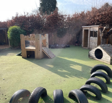 Wooden play fort sits on green turf, casting long shadows; nearby half-buried tires form a stepping course, with log tunnel and wooden shed before brown hedges under sunny sky.