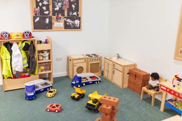 Wooden toy kitchen and shelving hold kids' safety vests and helmets while toy cars, trucks, stacked foam bricks and a seated doll scatter across a carpeted classroom corner.