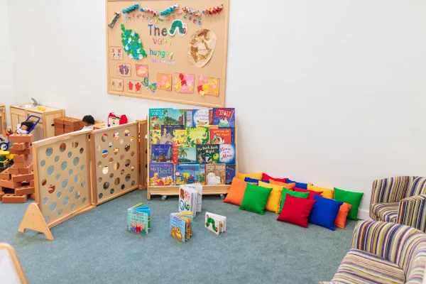Children's book rack filled with picture books; colorful floor cushions and striped armchairs arranged on carpet; a wooden play divider and toy blocks sit nearby in a bright classroom corner.