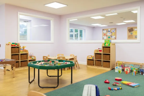 Low round activity table holds a wooden train set; surrounded by small chairs, toy shelves and cubbies, colorful toys on a carpeted corner inside a bright, pastel-painted preschool classroom.