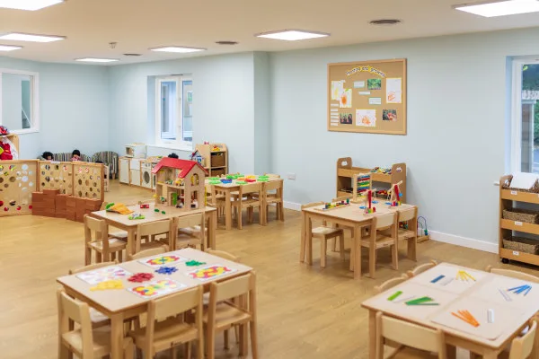Small wooden tables and chairs arranged and topped with colorful puzzles, blocks and craft supplies, occupying a bright, spacious preschool classroom with light walls, windows and wooden flooring.
